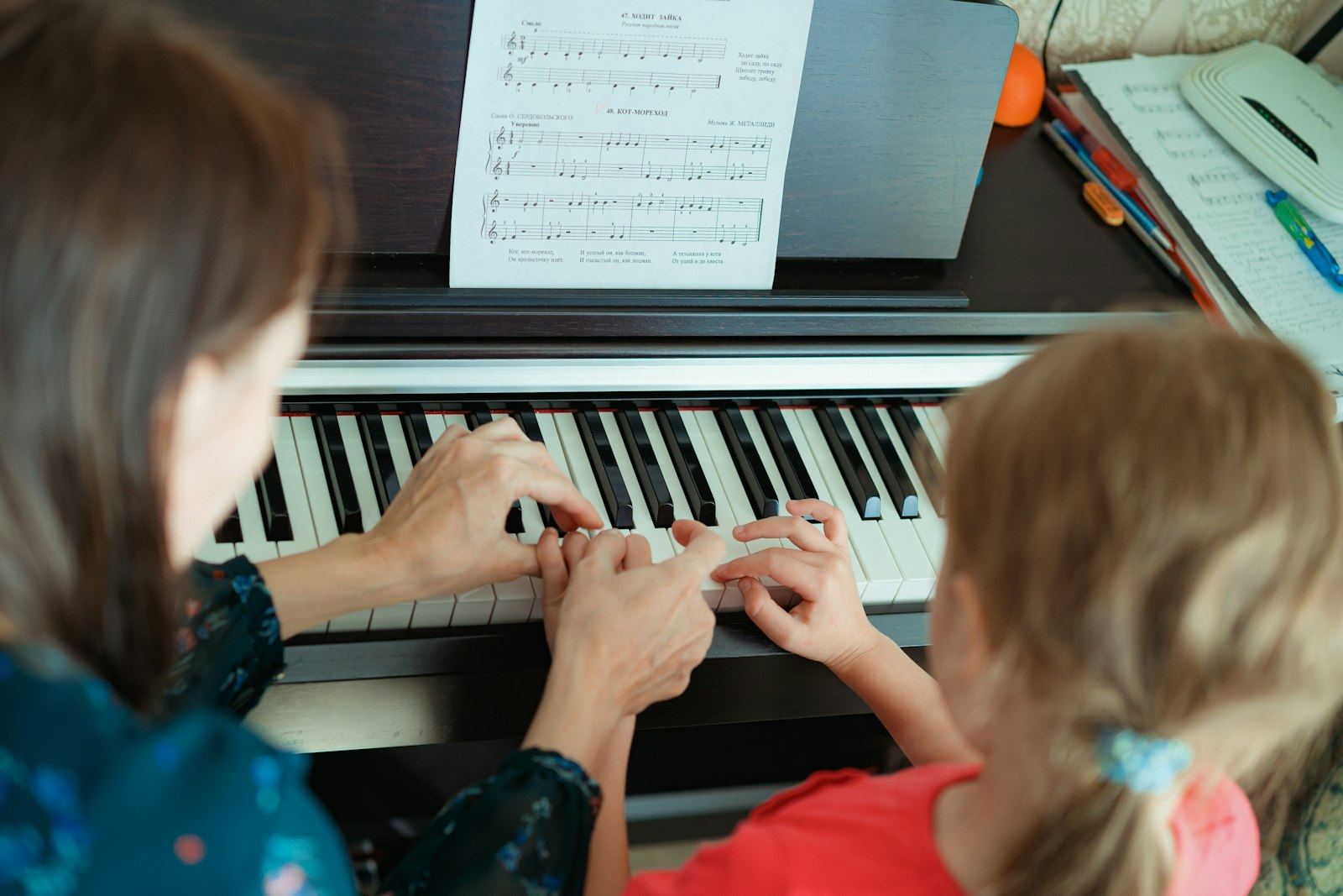 A teacher guides a young student's hands on the piano keys, with sheet music on the stand
